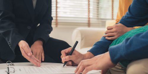 Happy young Asian couple and realtor agent. Cheerful young man signing some documents while sitting at desk together with his wife. Buying new house real estate. Signing good condition contract.